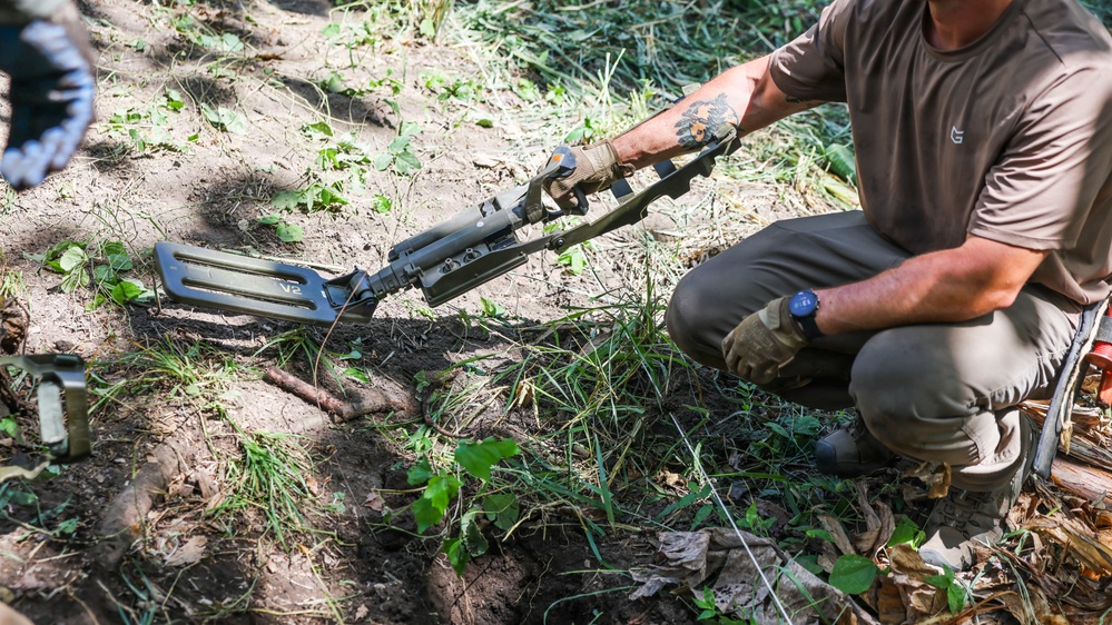 A person in tactical/outdoor clothing is crouching on the ground while operating what appears to be a metal detector or ground-scanning device labeled 'V2'. The individual is wearing gloves, a brown t-shirt, olive/khaki pants, and a watch, and has a visible tattoo on their forearm. The setting is an outdoor area with disturbed soil, grass, and vegetation.