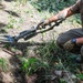 A person in tactical/outdoor clothing is crouching on the ground while operating what appears to be a metal detector or ground-scanning device labeled 'V2'. The individual is wearing gloves, a brown t-shirt, olive/khaki pants, and a watch, and has a visible tattoo on their forearm. The setting is an outdoor area with disturbed soil, grass, and vegetation.