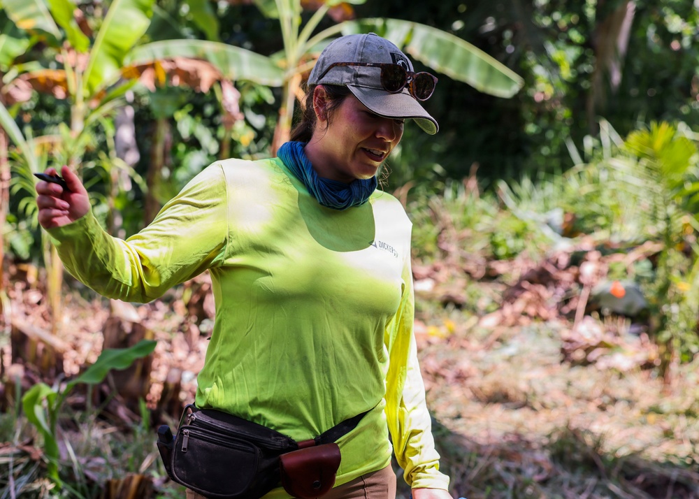 A woman is outdoors in a tropical or subtropical environment, dressed in field/outdoor work attire. She appears to be actively engaged in some form of fieldwork or guided activity, holding a small object in her right hand and speaking or gesturing. The lush green vegetation in the background, including banana plants, suggests a warm, humid climate.