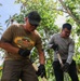 Two men are engaged in outdoor manual labor, likely digging or clearing ground, in a tropical setting surrounded by palm trees and lush vegetation. The man in the foreground wears a 'Beerlao' branded t-shirt, a dark cap, sunglasses, and black work gloves, and has a visible tattoo on his arm. Both individuals are bent forward and holding long-handled tools, suggesting active physical work.