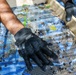 A person wearing black tactical/work gloves is using a wooden-framed wire mesh screen to sift soil and debris outdoors. The screen appears to be resting on a blue tarp, and the mesh contains dirt clumps, leaf fragments, and organic material. This activity is consistent with archaeological fieldwork, soil screening, or gardening/composting.
