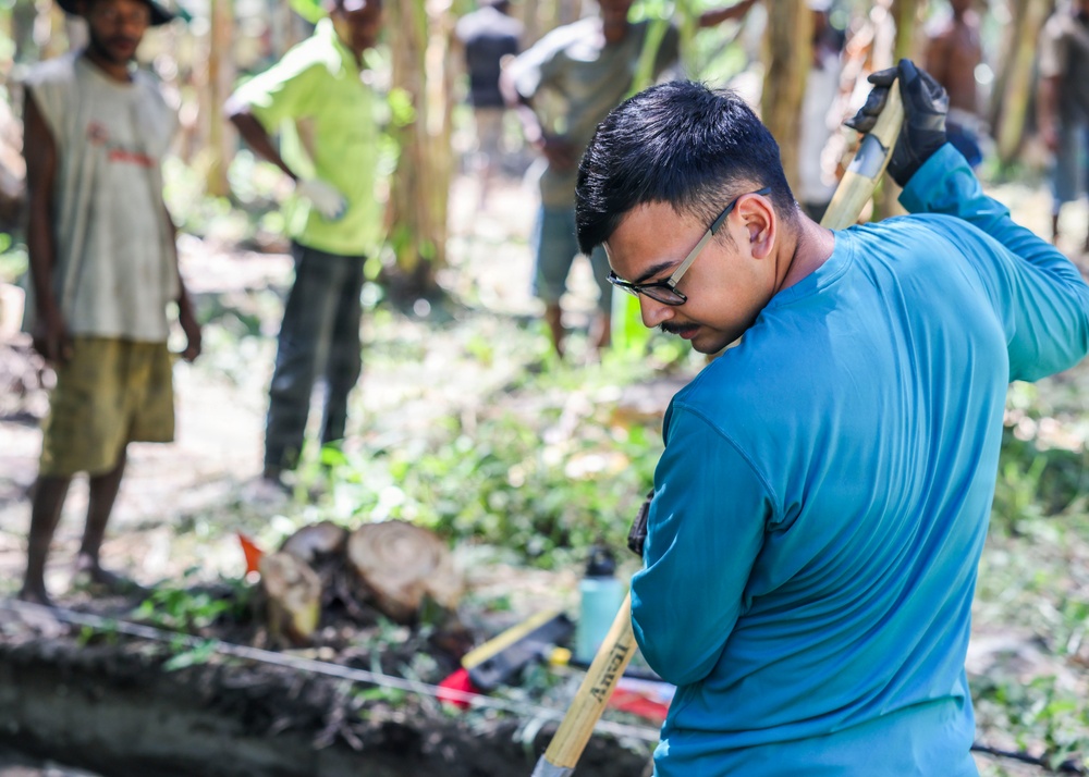 A young man wearing glasses and a teal/blue shirt is actively working with a long-handled tool, likely a shovel or hoe, in a forested outdoor setting. Several other individuals are visible in the blurred background, observing or participating in what appears to be a community work or volunteer activity. The scene suggests a land clearing, trail building, or community service project in a tropical or subtropical environment.