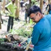 A young man wearing glasses and a teal/blue shirt is actively working with a long-handled tool, likely a shovel or hoe, in a forested outdoor setting. Several other individuals are visible in the blurred background, observing or participating in what appears to be a community work or volunteer activity. The scene suggests a land clearing, trail building, or community service project in a tropical or subtropical environment.