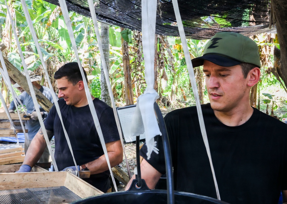 Two men in black t-shirts are working under a shade net structure in what appears to be a tropical outdoor setting surrounded by banana trees. They appear to be engaged in some form of manual labor or processing work involving wooden frames and black containers. A third person is partially visible in the background.
