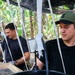 Two men in black t-shirts are working under a shade net structure in what appears to be a tropical outdoor setting surrounded by banana trees. They appear to be engaged in some form of manual labor or processing work involving wooden frames and black containers. A third person is partially visible in the background.