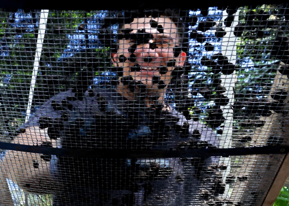 A smiling person is visible through a mesh or net screen, photographed from below or at an angle looking up through the netting. The dark mesh creates a grid-like overlay across the entire image, partially obscuring the subject. Green foliage and blue sky are visible in the background.