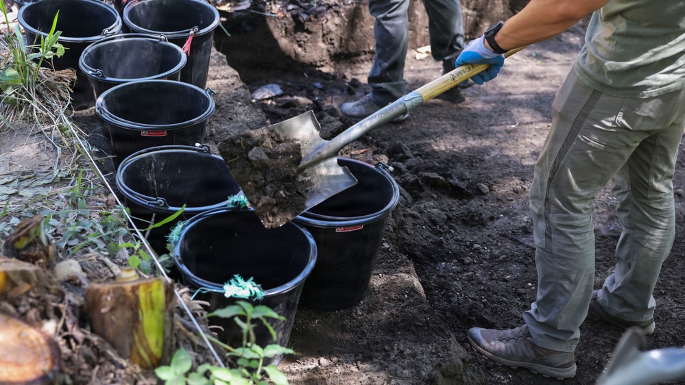 A worker wearing blue gloves and gray clothing is shoveling dark soil into black rubber buckets at what appears to be an excavation or archaeological dig site. Multiple black buckets are lined up along a string or rope boundary line, with at least one other person visible in the background. The setting is outdoors in a shaded, wooded area.