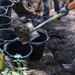A worker wearing blue gloves and gray clothing is shoveling dark soil into black rubber buckets at what appears to be an excavation or archaeological dig site. Multiple black buckets are lined up along a string or rope boundary line, with at least one other person visible in the background. The setting is outdoors in a shaded, wooded area.