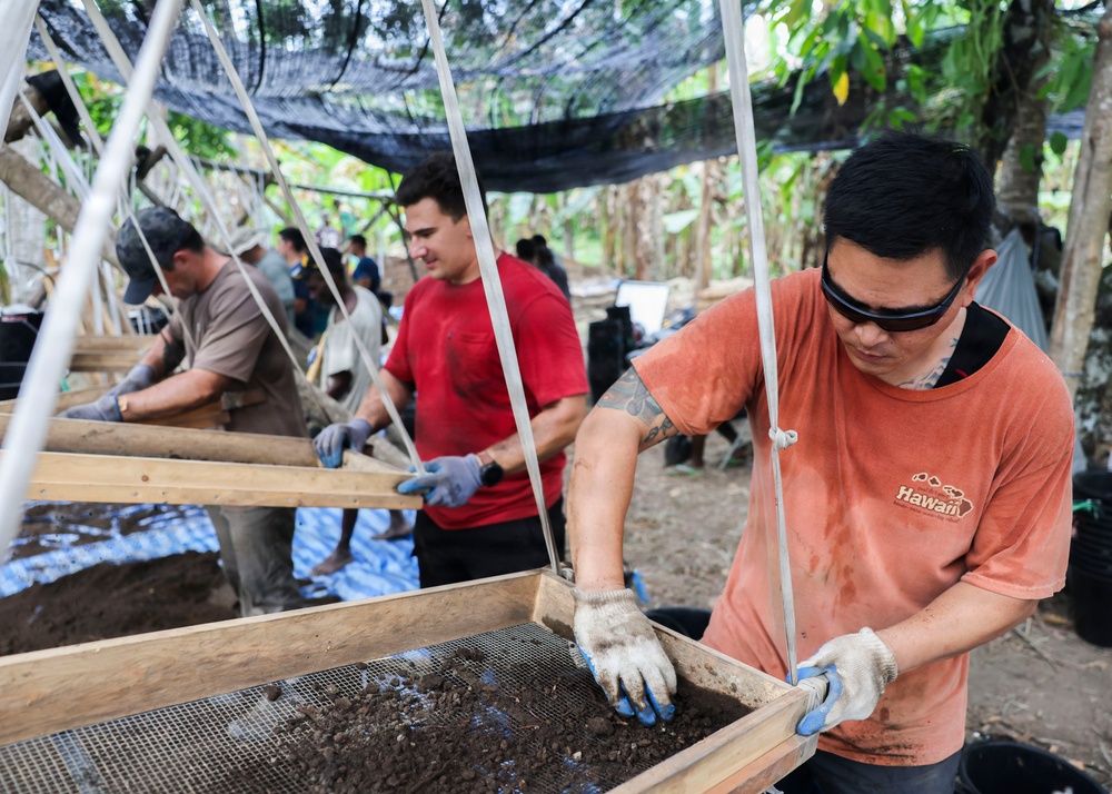 Multiple workers are engaged in an outdoor archaeological or soil-sifting operation under a shade canopy in what appears to be a tropical environment. The workers are using wooden-framed mesh screens to sift through soil and sediment, a common technique in archaeological excavations or forensic recovery operations. The setting, attire, and organized workflow suggest a coordinated field operation, possibly a military or humanitarian recovery mission.