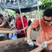 Multiple workers are engaged in an outdoor archaeological or soil-sifting operation under a shade canopy in what appears to be a tropical environment. The workers are using wooden-framed mesh screens to sift through soil and sediment, a common technique in archaeological excavations or forensic recovery operations. The setting, attire, and organized workflow suggest a coordinated field operation, possibly a military or humanitarian recovery mission.