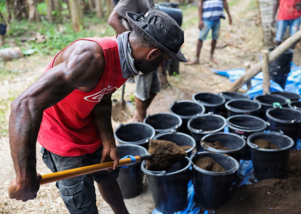 A man wearing a red Coca-Cola tank top and a dark wide-brimmed hat is shoveling soil or dirt into a row of black plastic buckets arranged on a blue tarp. Several other individuals are visible in the background in what appears to be an outdoor, forested or rural setting. The activity suggests agricultural preparation, possibly filling nursery pots with growing medium.