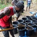 A man wearing a red Coca-Cola tank top and a dark wide-brimmed hat is shoveling soil or dirt into a row of black plastic buckets arranged on a blue tarp. Several other individuals are visible in the background in what appears to be an outdoor, forested or rural setting. The activity suggests agricultural preparation, possibly filling nursery pots with growing medium.