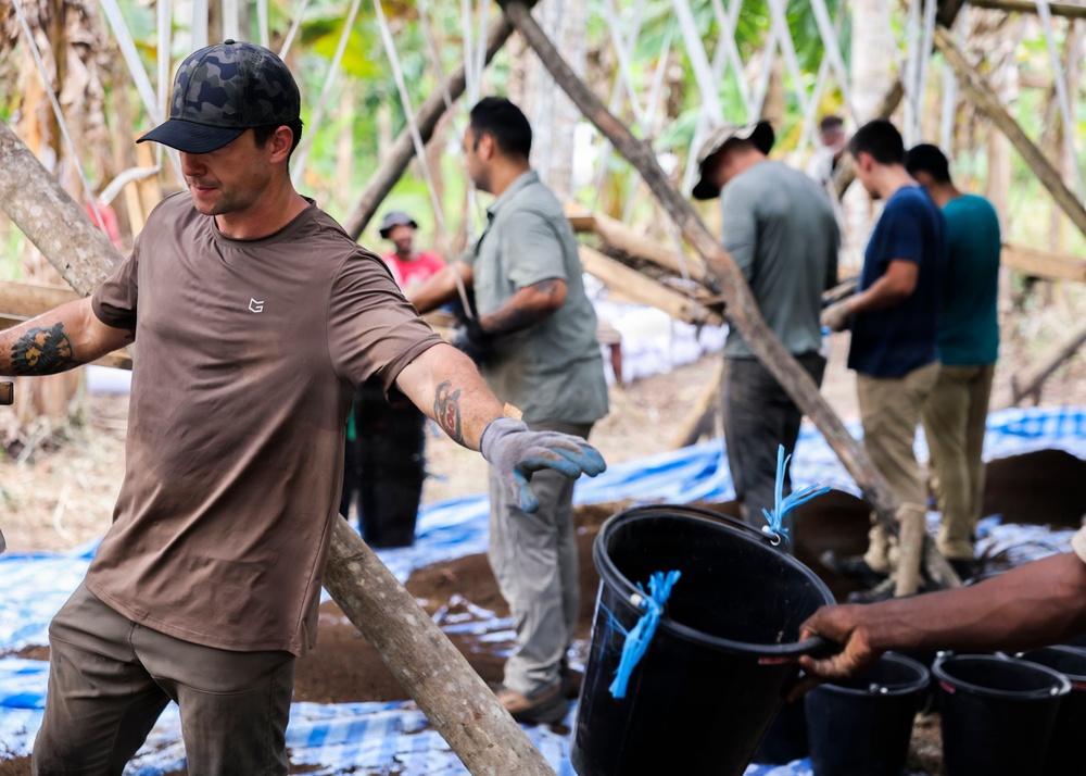 A group of workers are engaged in a construction or community project in a tropical outdoor setting. The foreground shows a tattooed man in a brown shirt and camo cap handling a wooden pole, while several others work in the background. Black buckets and blue tarps are visible on the ground, suggesting active manual labor.