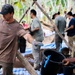 A group of workers are engaged in a construction or community project in a tropical outdoor setting. The foreground shows a tattooed man in a brown shirt and camo cap handling a wooden pole, while several others work in the background. Black buckets and blue tarps are visible on the ground, suggesting active manual labor.