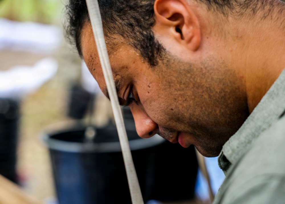 A close-up profile shot of a man with short dark curly hair and stubble, looking downward with visible sweat on his face. A diagonal strap or band crosses his face from forehead to chin. A dark bucket or container is visible in the blurred background.