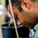 A close-up profile shot of a man with short dark curly hair and stubble, looking downward with visible sweat on his face. A diagonal strap or band crosses his face from forehead to chin. A dark bucket or container is visible in the blurred background.