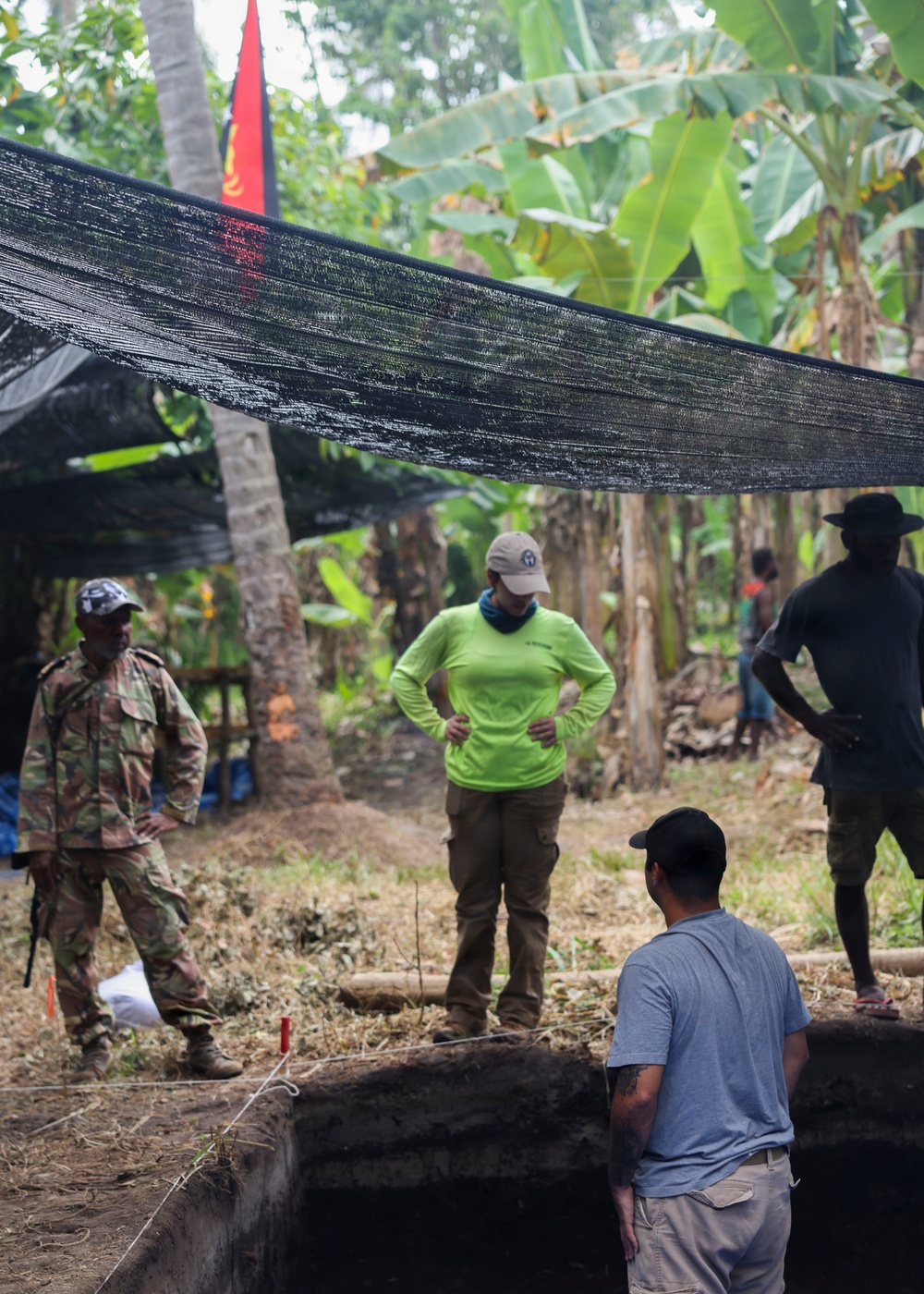 A field excavation site in a tropical environment, with several individuals gathered around a rectangular excavation pit. The team includes personnel in military camouflage, civilian field attire, and what appear to be local community members. A black shade net is stretched overhead, and a red and black flag is visible in the background among palm and banana trees.