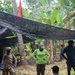 A field excavation site in a tropical environment, with several individuals gathered around a rectangular excavation pit. The team includes personnel in military camouflage, civilian field attire, and what appear to be local community members. A black shade net is stretched overhead, and a red and black flag is visible in the background among palm and banana trees.