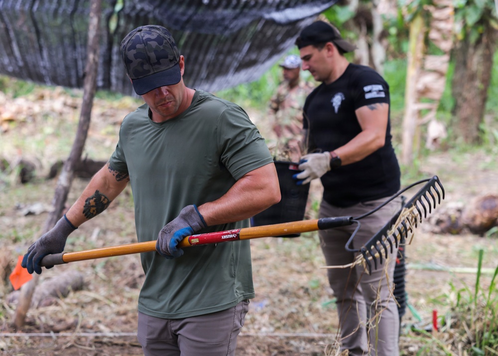Three individuals are engaged in outdoor manual labor, likely land clearing or site preparation in a tropical or subtropical environment. The foreground figure, wearing a green t-shirt and camo cap, is carrying a Husky-brand rake with visible tattoos on his arms. A second person in a black t-shirt with a patch and tactical pants works in the background alongside a third individual in camouflage attire.