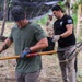 Three individuals are engaged in outdoor manual labor, likely land clearing or site preparation in a tropical or subtropical environment. The foreground figure, wearing a green t-shirt and camo cap, is carrying a Husky-brand rake with visible tattoos on his arms. A second person in a black t-shirt with a patch and tactical pants works in the background alongside a third individual in camouflage attire.