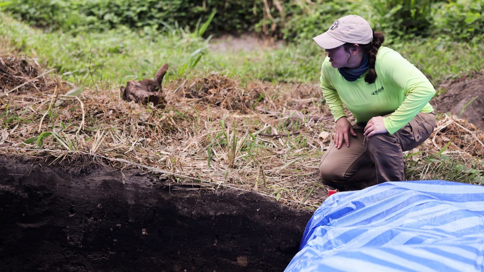 A field researcher or archaeologist crouches beside an excavation trench, closely examining the exposed soil profile. The dark, layered earth wall of the trench is clearly visible, suggesting deliberate stratigraphic excavation. A blue tarp is positioned at the edge of the pit, likely used to collect or protect excavated material.