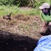 A field researcher or archaeologist crouches beside an excavation trench, closely examining the exposed soil profile. The dark, layered earth wall of the trench is clearly visible, suggesting deliberate stratigraphic excavation. A blue tarp is positioned at the edge of the pit, likely used to collect or protect excavated material.