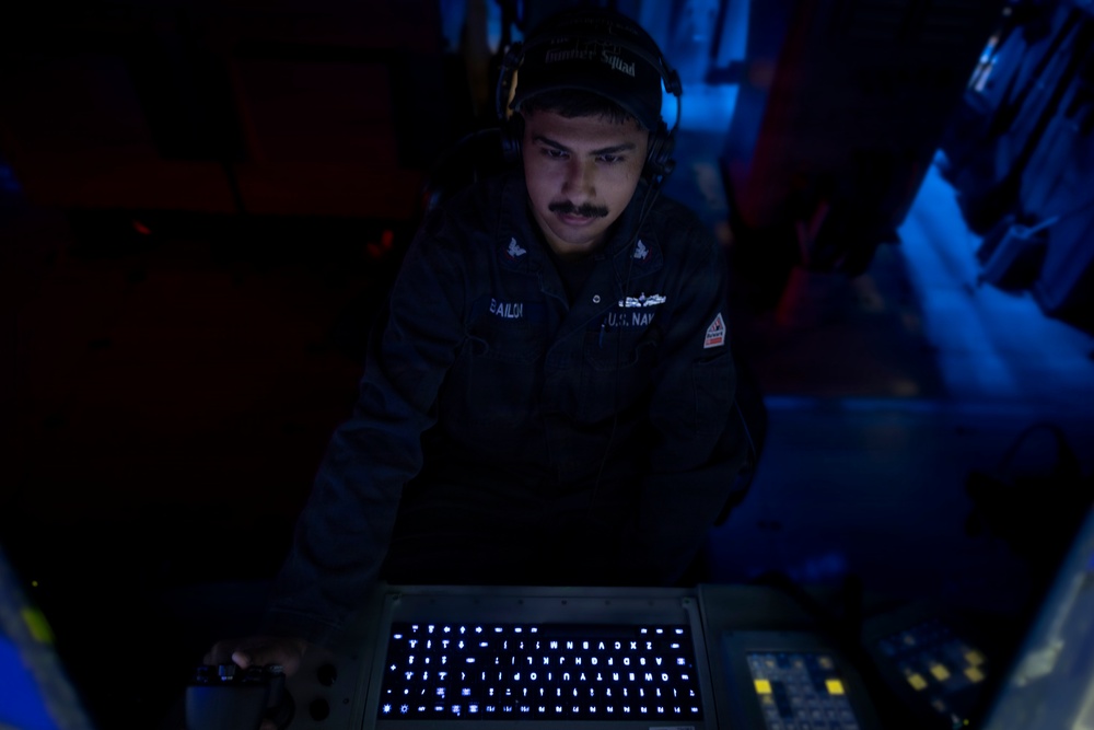 A U.S. Navy sailor wearing headphones and a dark uniform with 'U.S. NAVY' and a name tag reading 'GAILD_' is operating an illuminated keyboard/console in a dimly lit environment. The scene appears to be aboard a naval vessel, likely in a communications or operations station. The blue and red ambient lighting creates a dramatic, low-light operational atmosphere.