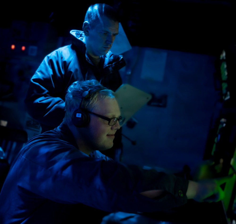 Two individuals are working in a dimly lit operations or command center, illuminated primarily by blue ambient light. The person in the foreground is seated at a workstation wearing headphones and glasses, while a second person stands behind observing the same display. The environment suggests a military or naval combat information center (CIC) based on the equipment and lighting conditions.