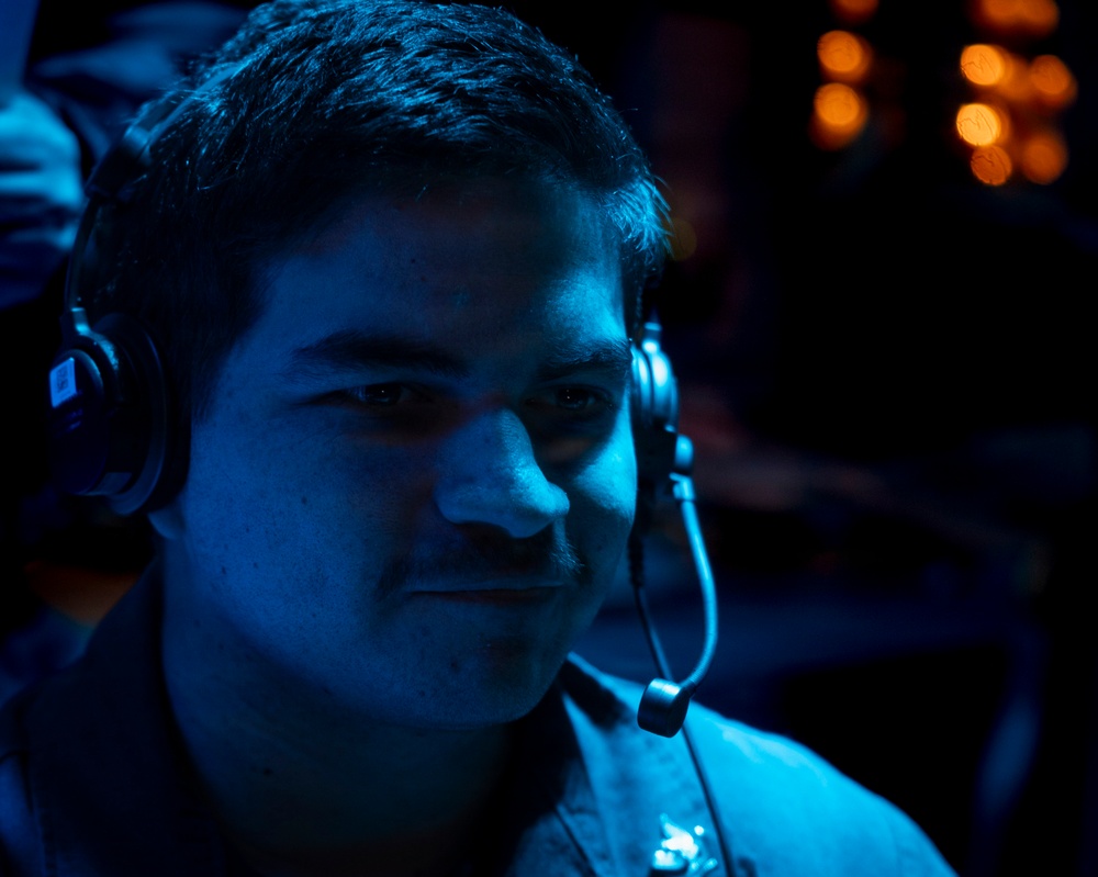 A young male is photographed in a dimly lit environment illuminated predominantly by blue ambient lighting. He is wearing a gaming or communications headset with a boom microphone positioned near his mouth. The background contains out-of-focus warm orange bokeh lights, suggesting a large indoor venue such as an esports event or gaming convention.