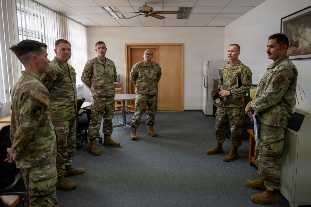 Six military personnel in OCP (Operational Camouflage Pattern) uniforms are gathered in a circle inside an office or administrative room. They appear to be engaged in a briefing, discussion, or informal meeting. The setting suggests a military installation building, likely in Europe based on the architectural style.