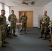 Six military personnel in OCP (Operational Camouflage Pattern) uniforms are gathered in a circle inside an office or administrative room. They appear to be engaged in a briefing, discussion, or informal meeting. The setting suggests a military installation building, likely in Europe based on the architectural style.
