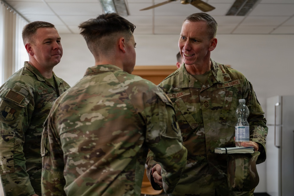 Three military personnel in OCP (Operational Camouflage Pattern) uniforms are engaged in conversation in what appears to be an indoor facility. The individual on the right, whose name tape reads 'CLARK' and whose uniform is marked 'U.S. AIR FORCE,' is speaking with a service member whose back is to the camera. A third service member stands in the background observing the interaction.