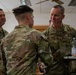 Three military personnel in OCP (Operational Camouflage Pattern) uniforms are engaged in conversation in what appears to be an indoor facility. The individual on the right, whose name tape reads 'CLARK' and whose uniform is marked 'U.S. AIR FORCE,' is speaking with a service member whose back is to the camera. A third service member stands in the background observing the interaction.