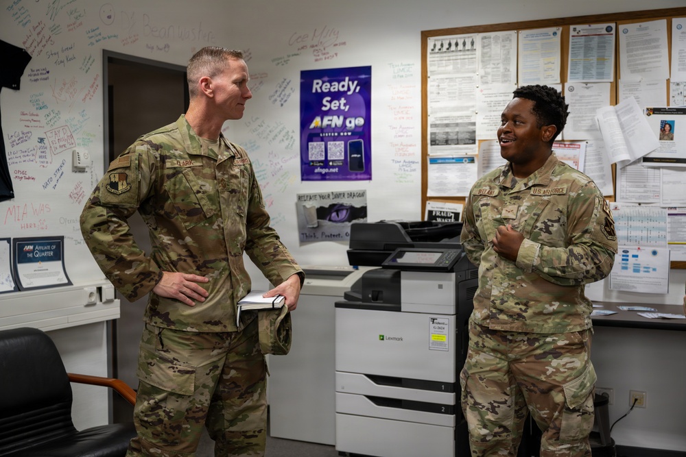 Two U.S. Air Force personnel in OCP (Operational Camouflage Pattern) uniforms are engaged in conversation inside what appears to be a military office or workspace. The setting includes a bulletin board with posted notices, a Lexmark printer/copier, and a whiteboard covered in handwritten notes and signatures. An AFN (American Forces Network) promotional poster is visible in the background.