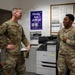 Two U.S. Air Force personnel in OCP (Operational Camouflage Pattern) uniforms are engaged in conversation inside what appears to be a military office or workspace. The setting includes a bulletin board with posted notices, a Lexmark printer/copier, and a whiteboard covered in handwritten notes and signatures. An AFN (American Forces Network) promotional poster is visible in the background.