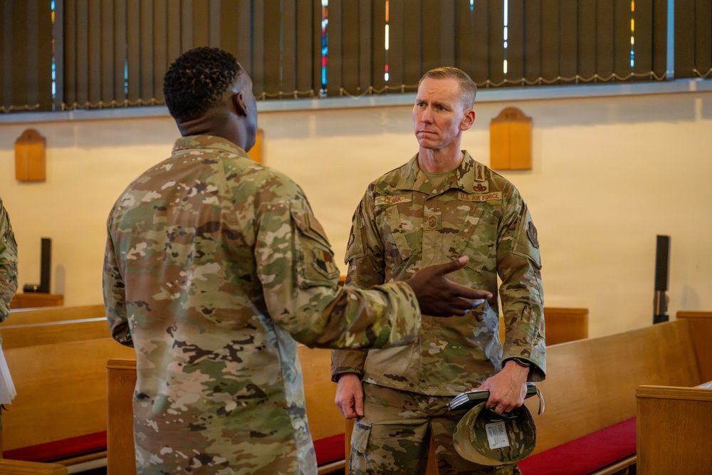 Two U.S. Air Force personnel in OCP (Operational Camouflage Pattern) uniforms are engaged in conversation inside what appears to be a military chapel. The servicemember facing the camera has a name tape partially readable as 'CLARK' and 'U.S. AIR FORCE' tape visible, and is holding a patrol cap in his hand. The setting features wooden pews with red cushions and stained glass windows, consistent with a military base chapel.
