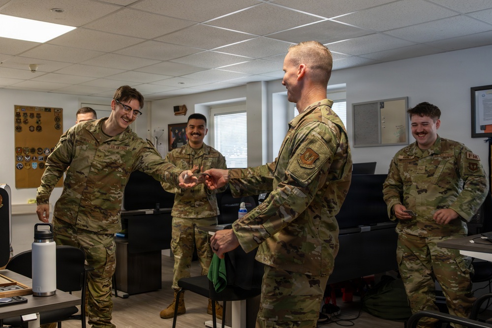 A group of U.S. Air Force personnel in OCP (Operational Camouflage Pattern) uniforms are gathered in what appears to be an office or briefing room. A senior airman appears to be presenting or receiving a coin or small object from another service member, a common military tradition known as a 'coin check' or coin presentation ceremony. The atmosphere is relaxed and jovial, with multiple personnel smiling and laughing.
