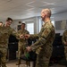 A group of U.S. Air Force personnel in OCP (Operational Camouflage Pattern) uniforms are gathered in what appears to be an office or briefing room. A senior airman appears to be presenting or receiving a coin or small object from another service member, a common military tradition known as a 'coin check' or coin presentation ceremony. The atmosphere is relaxed and jovial, with multiple personnel smiling and laughing.