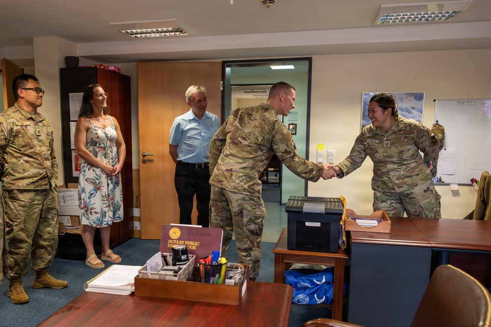 A military office setting where two uniformed service members in U.S. Army/Air Force OCP (Operational Camouflage Pattern) uniforms are shaking hands across a desk. Three civilians and another service member observe the interaction, suggesting a formal recognition or greeting ceremony. A 'Notary Public Record Book' is visible on the desk, indicating an administrative or legal services office.