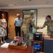 A military office setting where two uniformed service members in U.S. Army/Air Force OCP (Operational Camouflage Pattern) uniforms are shaking hands across a desk. Three civilians and another service member observe the interaction, suggesting a formal recognition or greeting ceremony. A 'Notary Public Record Book' is visible on the desk, indicating an administrative or legal services office.