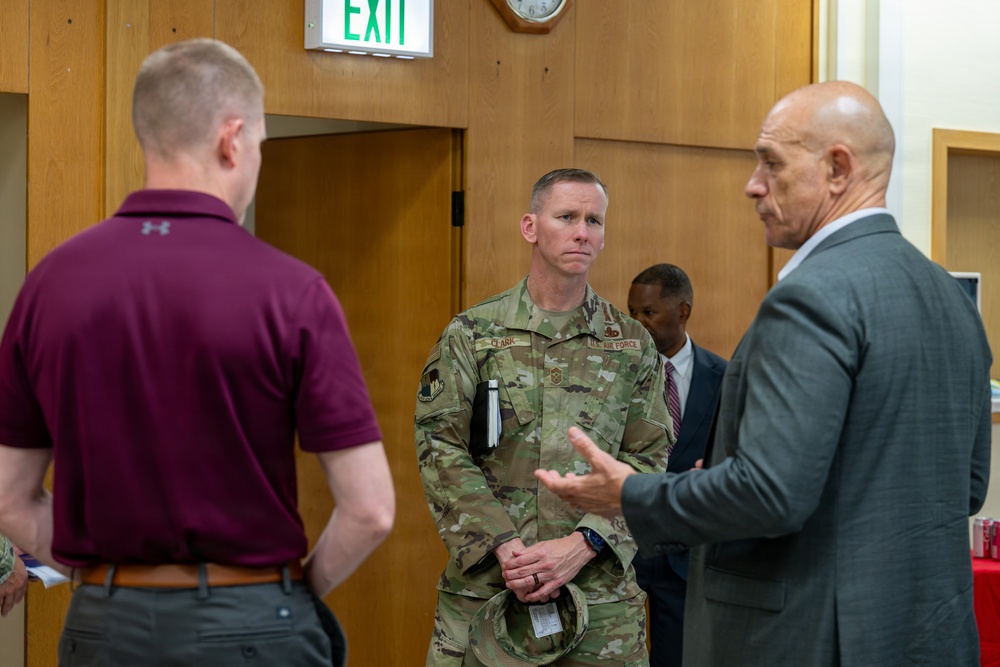 A group of individuals are engaged in conversation in what appears to be an indoor facility. A U.S. Air Force service member in OCP (Operational Camouflage Pattern) uniform, identified by a name tape reading 'CLARK,' is listening attentively while a man in a gray suit gestures as he speaks. A fourth person is visible in the background wearing a dark suit.