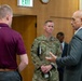 A group of individuals are engaged in conversation in what appears to be an indoor facility. A U.S. Air Force service member in OCP (Operational Camouflage Pattern) uniform, identified by a name tape reading 'CLARK,' is listening attentively while a man in a gray suit gestures as he speaks. A fourth person is visible in the background wearing a dark suit.