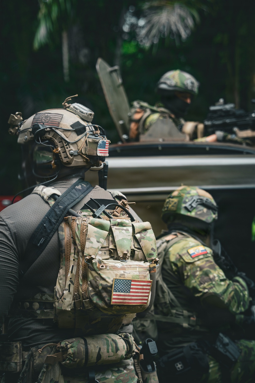 The image shows multiple heavily equipped military or special operations personnel in a tropical/jungle environment. At least three individuals are visible, wearing full tactical gear including helmets, body armor, and carrying weapons. Two distinct national flag patches are visible: United States and what appears to be Ecuador or Colombia.