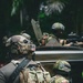 The image shows multiple heavily equipped military or special operations personnel in a tropical/jungle environment. At least three individuals are visible, wearing full tactical gear including helmets, body armor, and carrying weapons. Two distinct national flag patches are visible: United States and what appears to be Ecuador or Colombia.