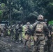 A group of armed military or law enforcement personnel in tactical gear are assembled on a muddy jungle path surrounded by dense tropical vegetation. Multiple operators are visible wearing camouflage uniforms, tactical vests, helmets, and carrying firearms. At least two individuals display visible US flag patches on their equipment.