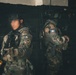Two armed military or special operations personnel are positioned against a dark wall, wearing camouflage uniforms and tactical gear. One individual's face has been deliberately blurred for anonymity. The soldier on the right displays what appears to be a flag patch on their sleeve, and both carry weapons and tactical equipment.