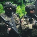 The image shows a group of heavily armed military or special operations personnel in a jungle or forested environment. Personnel are wearing multicam camouflage uniforms, tactical helmets, and face coverings, and are carrying assault rifles. Visible patches include Ecuadorian flag insignia and a US flag patch, suggesting a joint or multinational operation.