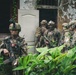 The image shows a group of heavily armed and equipped military personnel conducting what appears to be a building clearance or entry operation in a jungle or tropical environment. At least two nationalities are visibly represented, with one soldier displaying a Peruvian flag patch and another displaying a U.S. flag patch, suggesting a joint military exercise or operation. The soldiers are positioned around a deteriorating structure with an open doorway, surrounded by dense tropical vegetation.