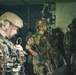 The image shows a group of military personnel in tactical gear inside a deteriorated building, likely during a joint training exercise or operation. At least two nationalities are identifiable: one soldier bears a US flag patch and another displays what appears to be a Peruvian flag patch. The personnel are equipped with helmets, body armor, weapons, and various tactical accessories.