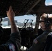 Two military aviators are seated in the cockpit of a large military aircraft, likely a transport or bomber, conducting pre-flight or ground operations checks. The pilot on the right wears a Bose headset and a flight suit with a visible U.S. flag patch, while the person on the left reaches up to manipulate overhead panel switches or controls. The aircraft appears to be on a flight line or tarmac, with a hangar visible through the cockpit windows.